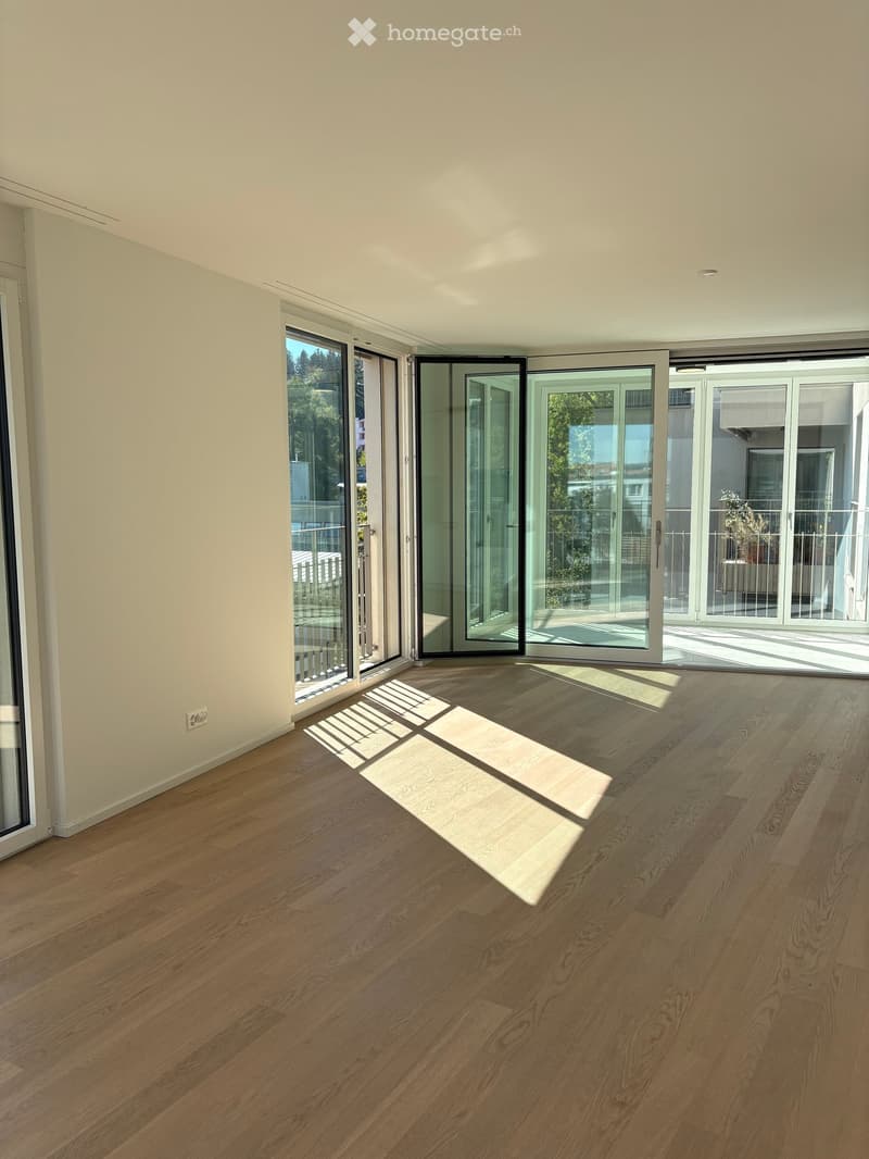 Empty room with light wooden flooring, large glass sliding doors leading to a balcony, and a small white rug on the floor.
