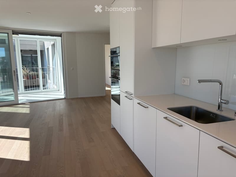 Modern kitchen with white cabinets, light wooden flooring, and a sink with a modern faucet. An oven is visible, and a sliding glass door leads to a balcony.