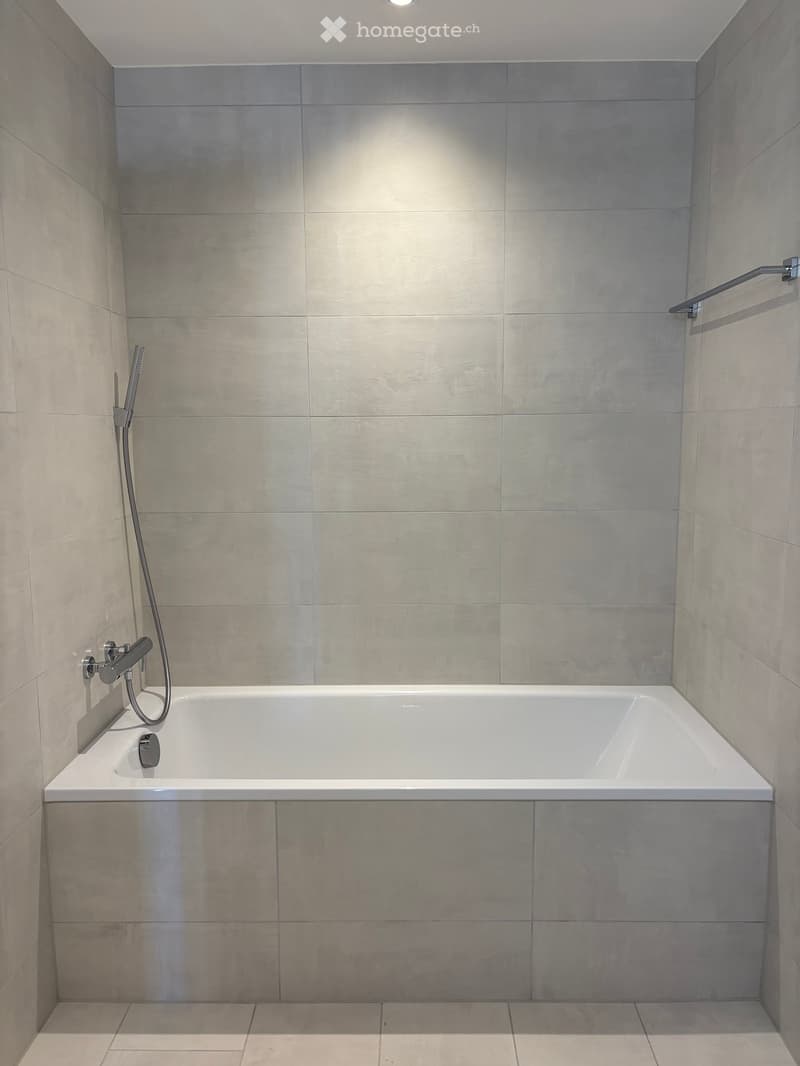Modern bathroom with a white bathtub, grey tiled walls, and a handheld showerhead attached to the wall.