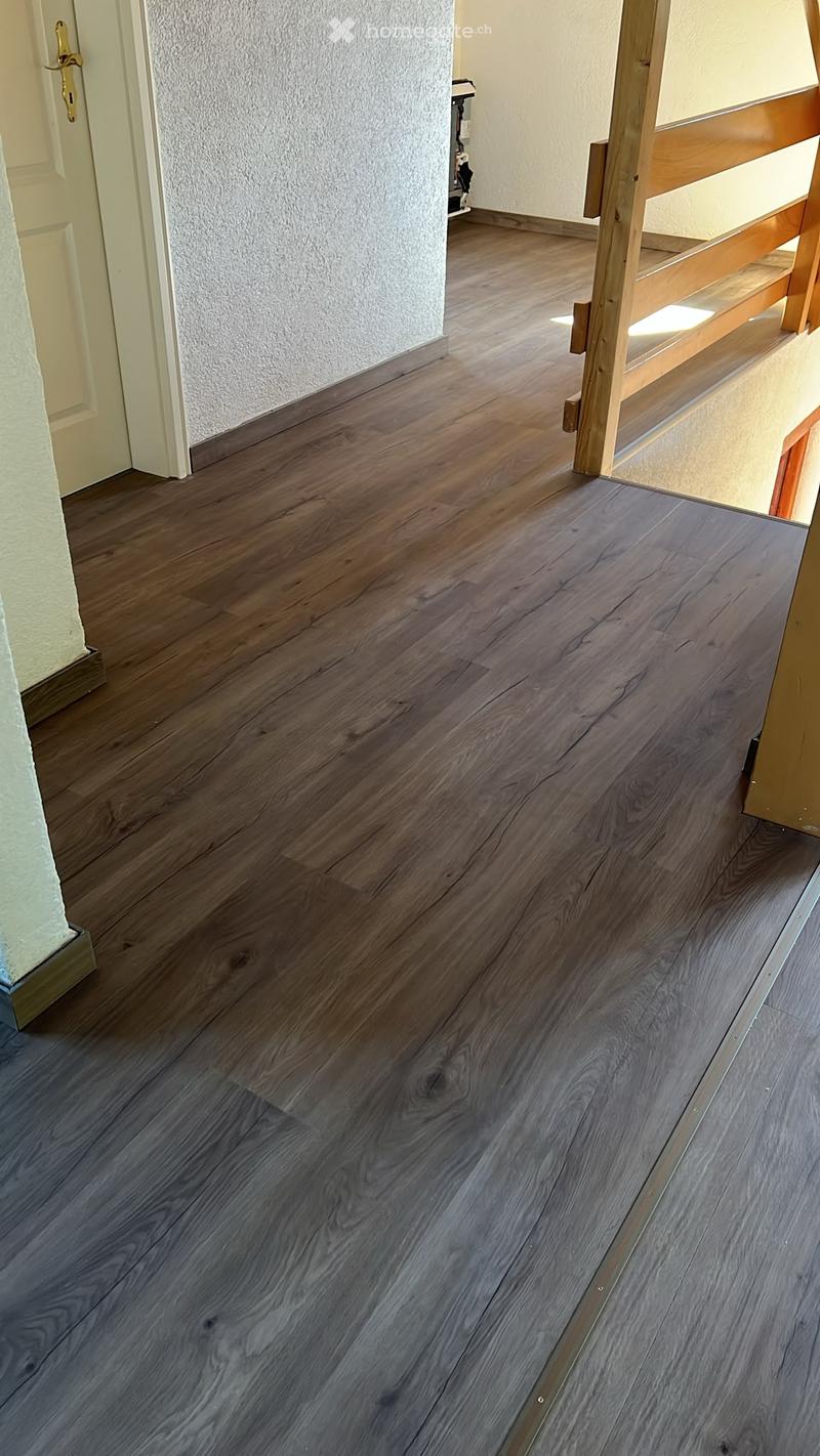 A hallway with dark wooden flooring and white walls, featuring a partially visible door frame and a wooden staircase in the background.