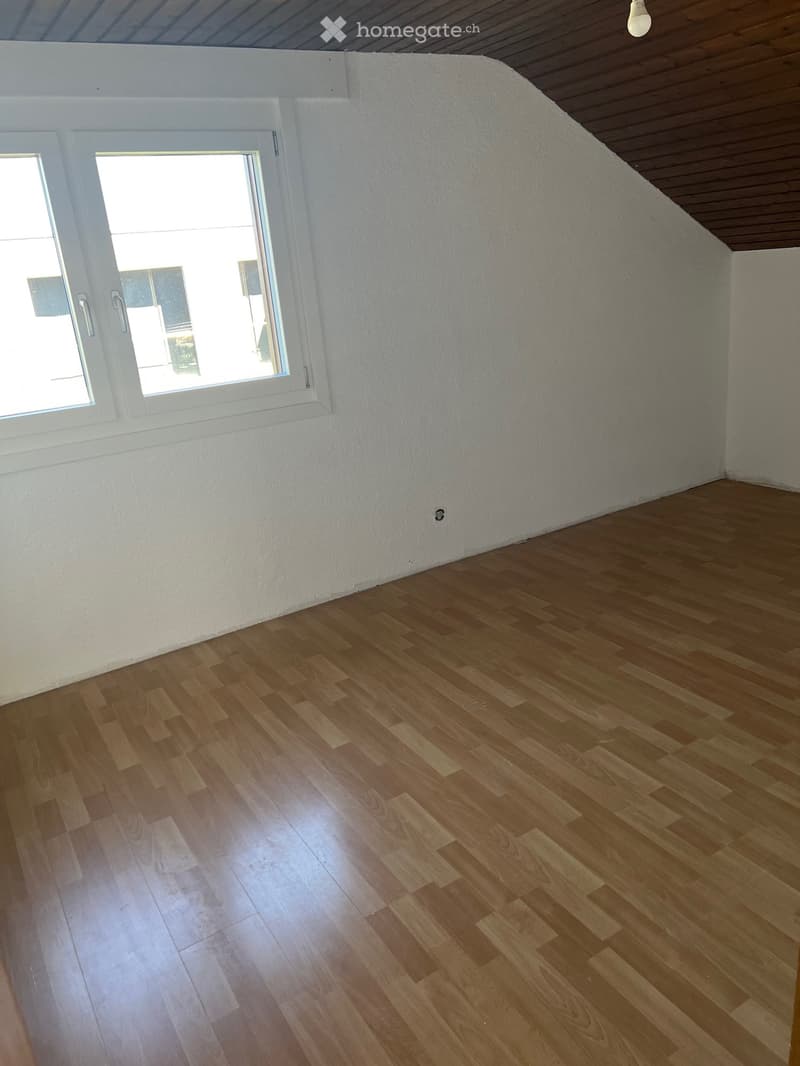 Empty room with wooden flooring and white walls, featuring a large window with white frame and a slanted ceiling with wooden beams.