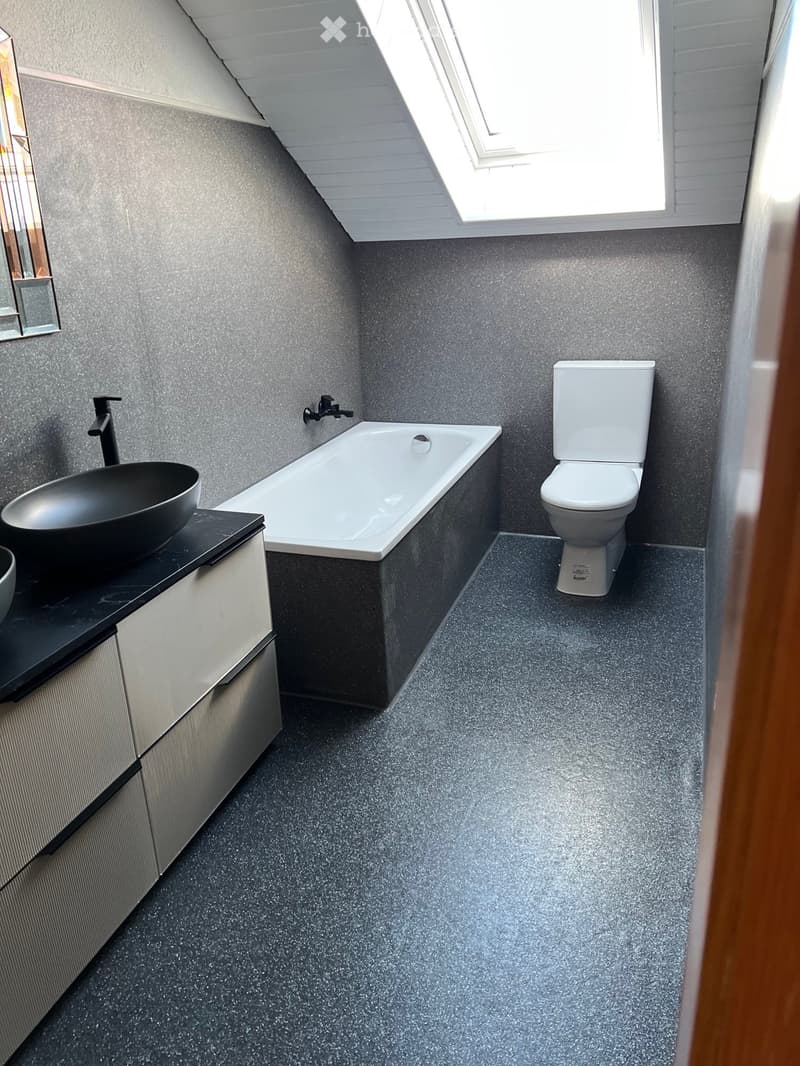 Modern bathroom with grey walls, a bathtub, toilet, sink, and a sleek black faucet. Features a skylight window for natural light.