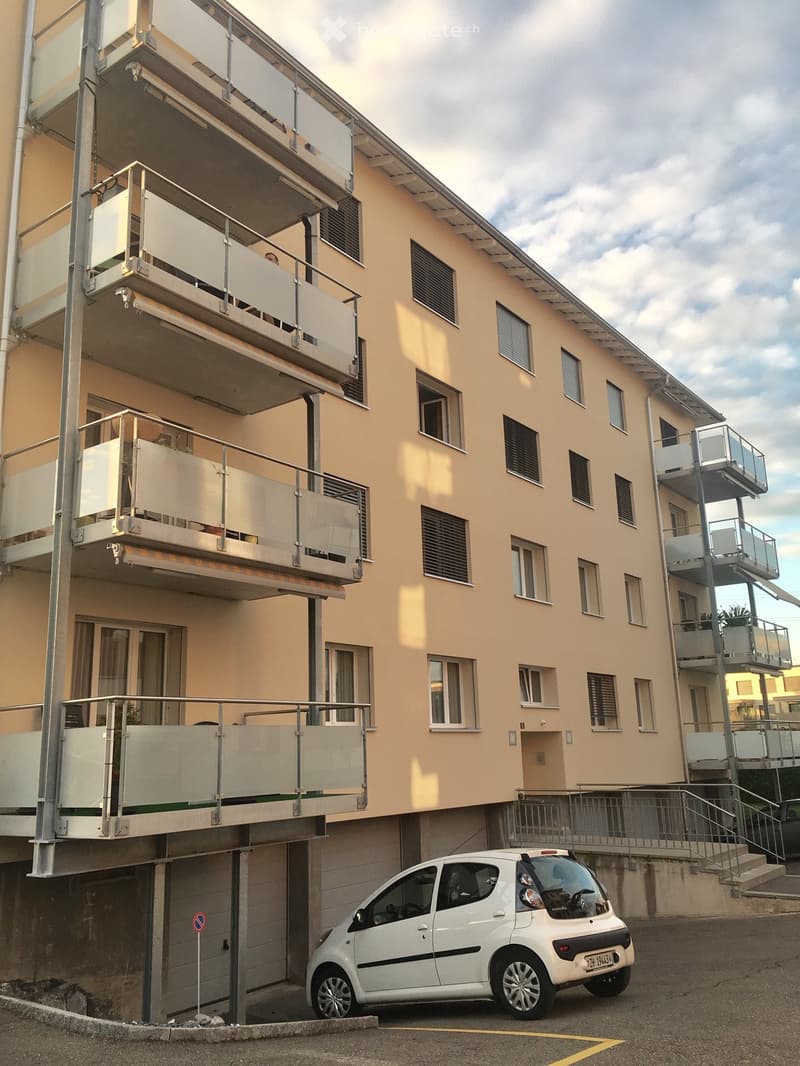 Multi-story residential building with beige facade, multiple windows, and several balconies with glass railings. A small white car is parked in the driveway.
