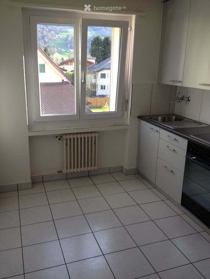 Empty kitchen with white tiled floor, white cabinets, sink, oven, and radiator under large windows overlooking a green landscape.