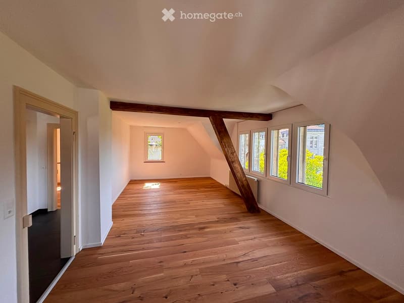 Empty attic room with wooden flooring, exposed wooden beams, and large windows allowing natural light. The space is unfurnished and has a sloped ceiling.