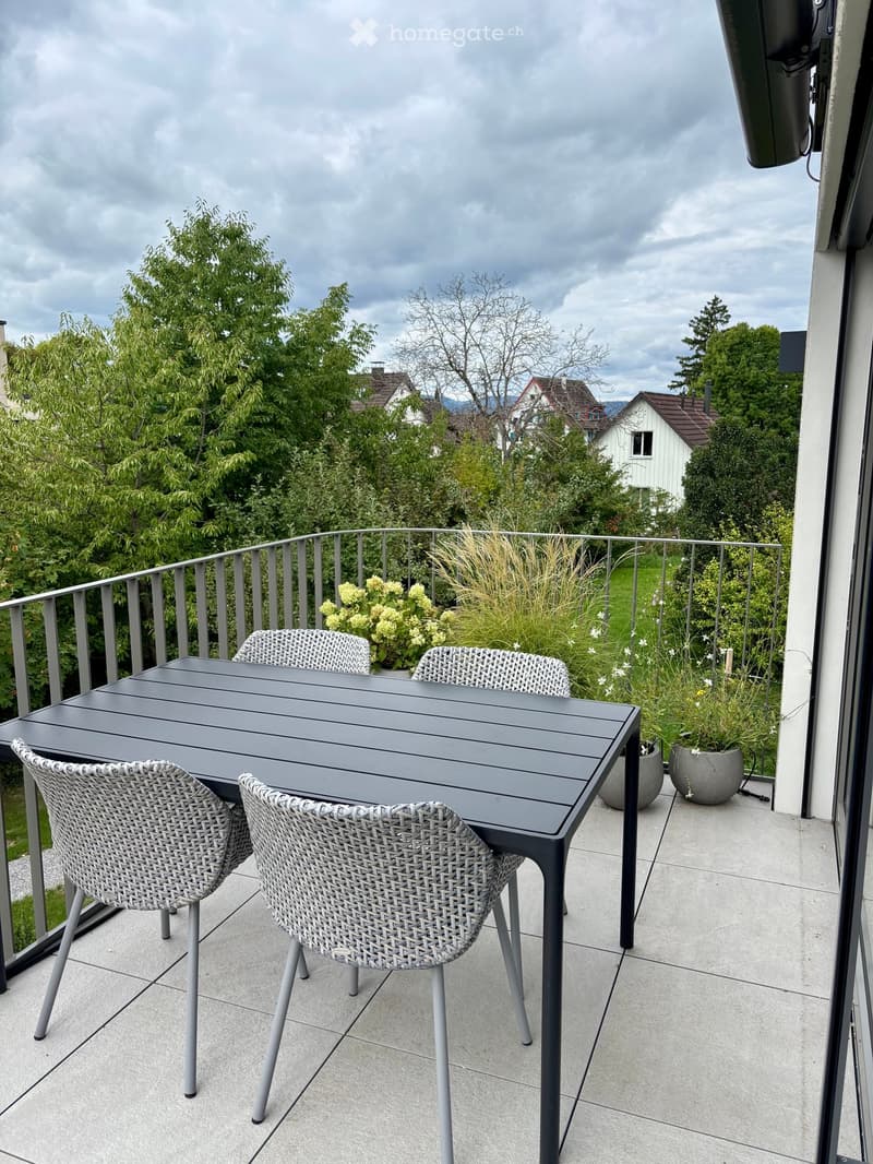 Balcony with a grey table and four wicker chairs, surrounded by potted plants and a metal railing, overlooking a green landscape with trees and houses.