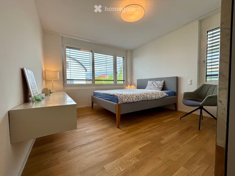 Modern bedroom with wooden flooring, large windows with blinds, a grey bed with a patterned duvet, a grey armchair, a side table with a lamp, and a picture frame on a console table.