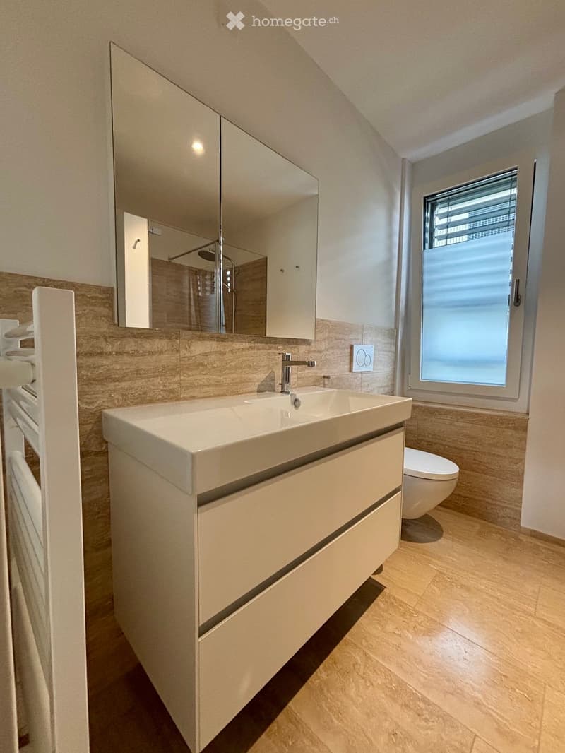 Modern bathroom with a large sink, mirror, toilet, and a window with blinds. The walls are tiled in a light, neutral color, and the floor is made of light-colored tiles.