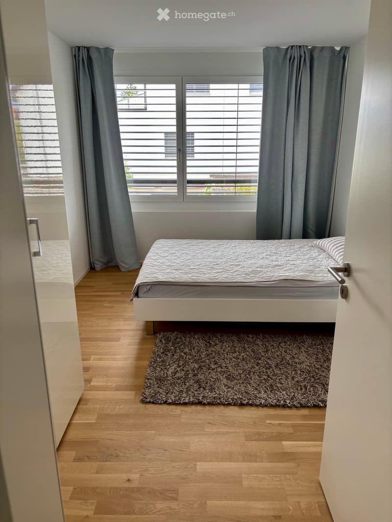 Minimalist bedroom with a single bed, grey bedding, a small shaggy rug, and large windows with grey curtains.