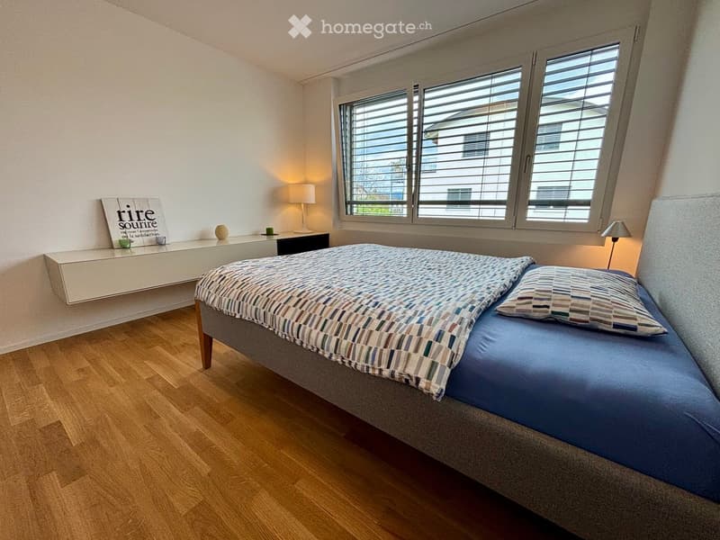 Modern bedroom with wooden flooring, large window with blinds, grey upholstered bed with patterned bedding, nightstand with lamp, decorative vase, and a book on the shelf.