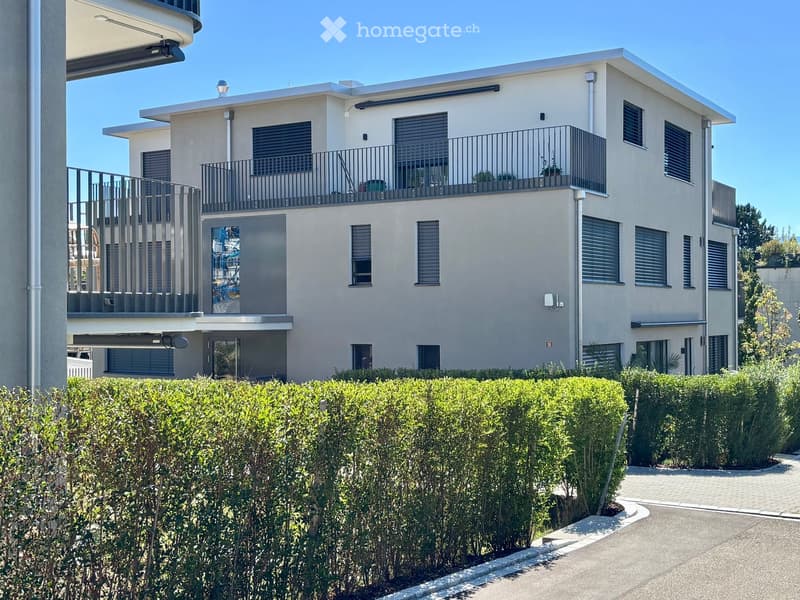 Modern two-story house with light beige exterior, multiple windows with shutters, balconies on both levels, and a well-maintained hedge in the front yard.