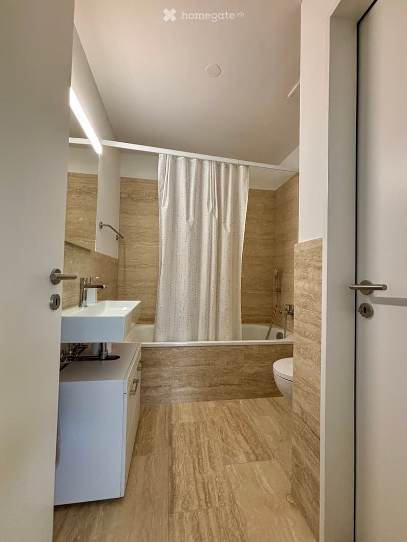 Modern bathroom with light wood flooring, a white sink with a chrome faucet, a bathtub with a shower curtain, and a toilet. The walls are tiled in a light wood pattern.