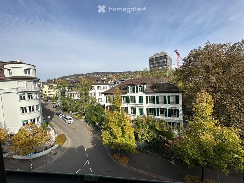 Urban street view, multi-story buildings, parked cars, trees, distant construction site