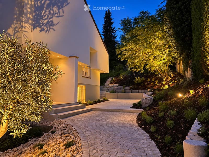 Modern two-story house with a raised ground floor, illuminated by warm lighting, surrounded by landscaped garden with steps and pathways.