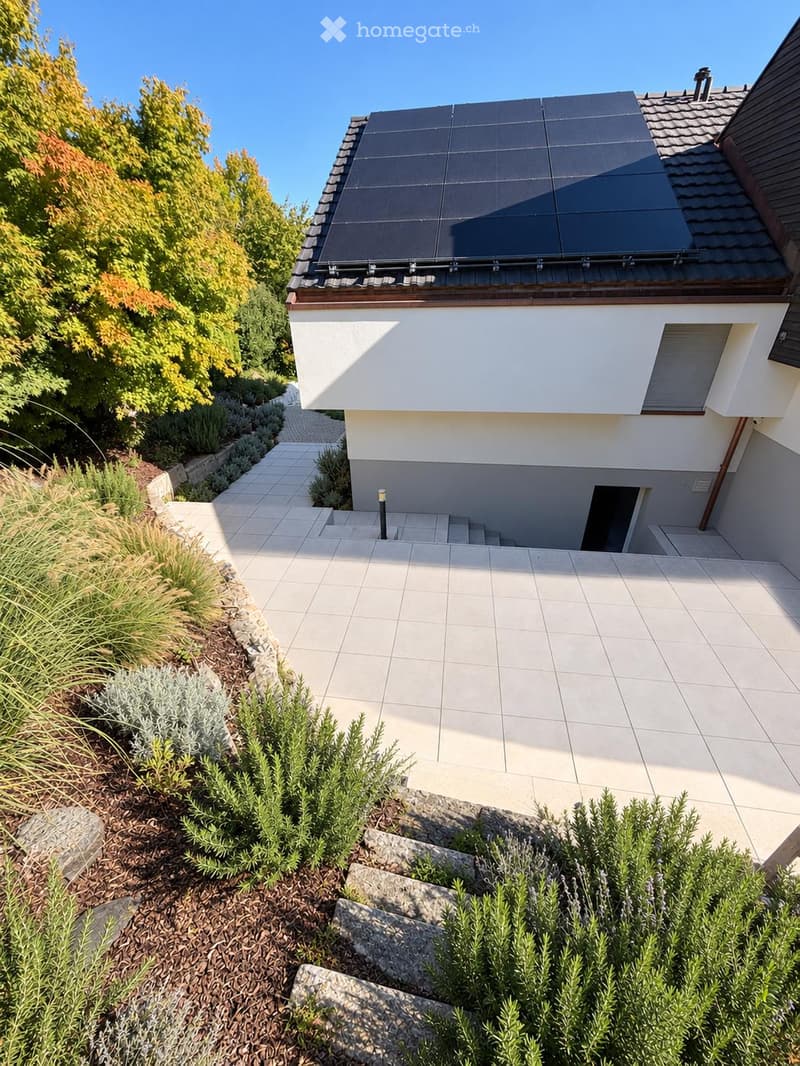 Modern house with solar panels on the roof, surrounded by a well-maintained garden with shrubs, plants, and a paved pathway leading to the entrance.