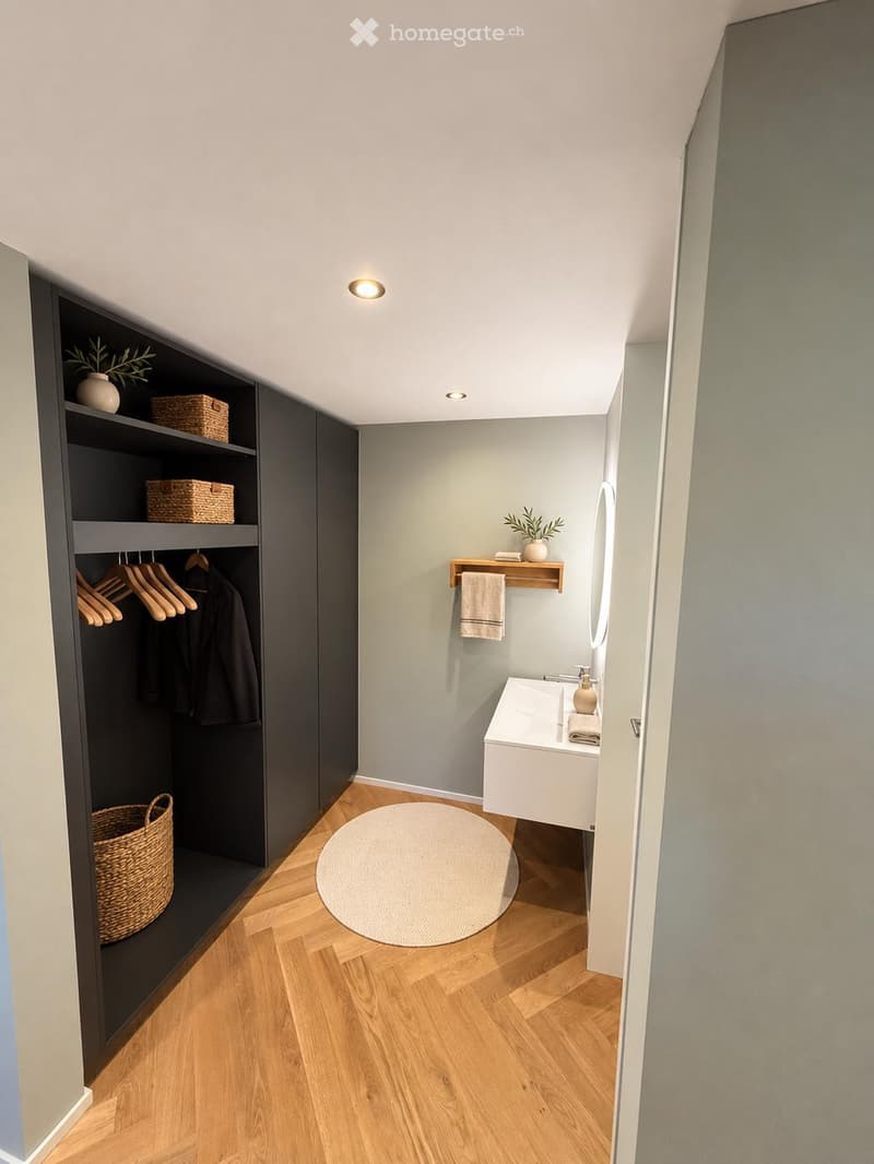 Modern bathroom with light grey walls, wooden flooring, a white sink, a round rug, a built-in dark cabinet with hanging clothes, and decorative baskets.