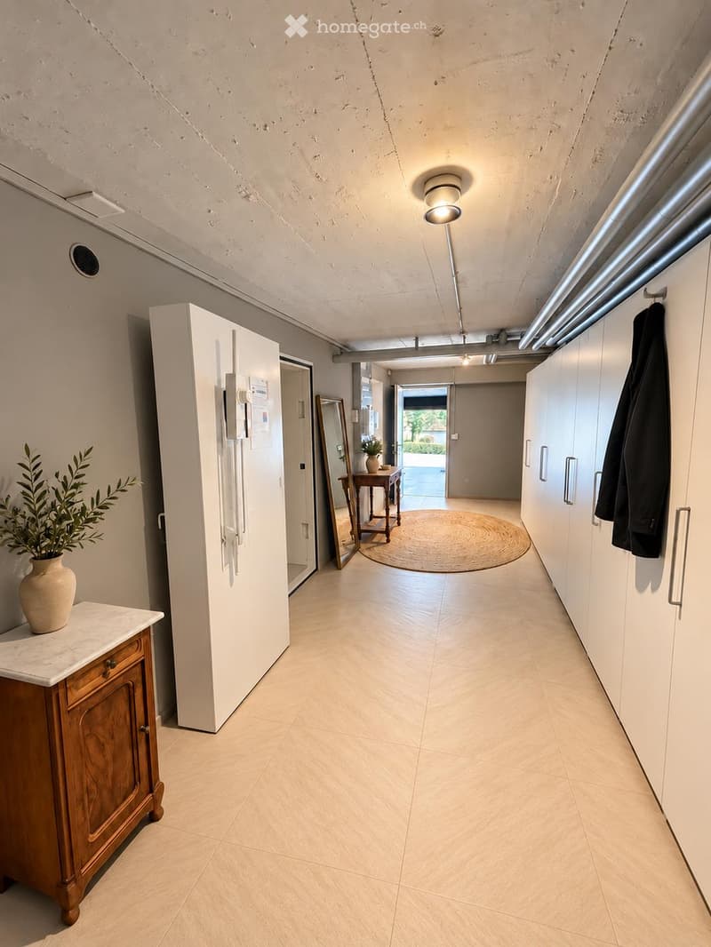 Modern hallway with light-colored tiles, white walls, built-in storage units, and a small wooden side table with a plant. A coat is hanging on the right side, and a round rug is visible in the adjacent room.