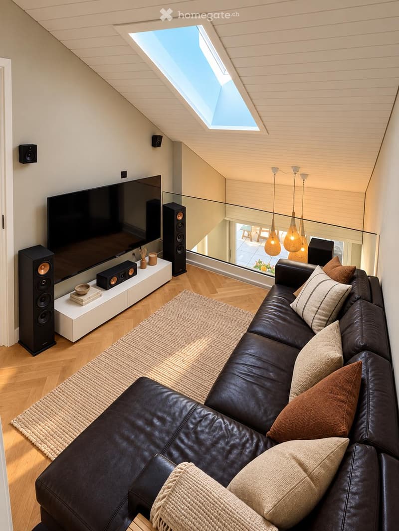 Modern living room with a large black leather sectional sofa, multiple throw pillows, a woven area rug, a sleek white coffee table holding books and a bowl, and a large flat-screen TV mounted on the wall with speakers. The room features wooden flooring, a