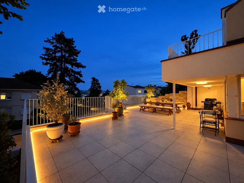 Well-lit outdoor patio with tiled flooring, potted plants, a wooden bench, and a grill, surrounded by a white railing and trees.