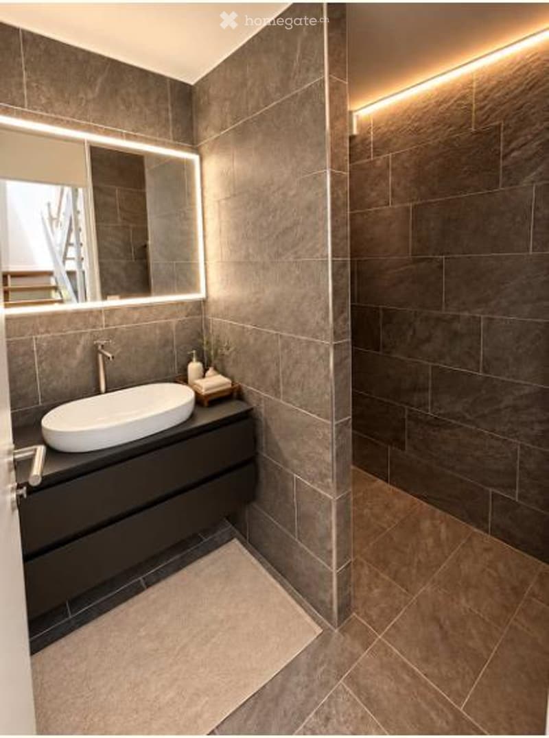 Modern bathroom with dark grey tiles, a white sink, a large illuminated mirror, and a sleek black vanity unit.