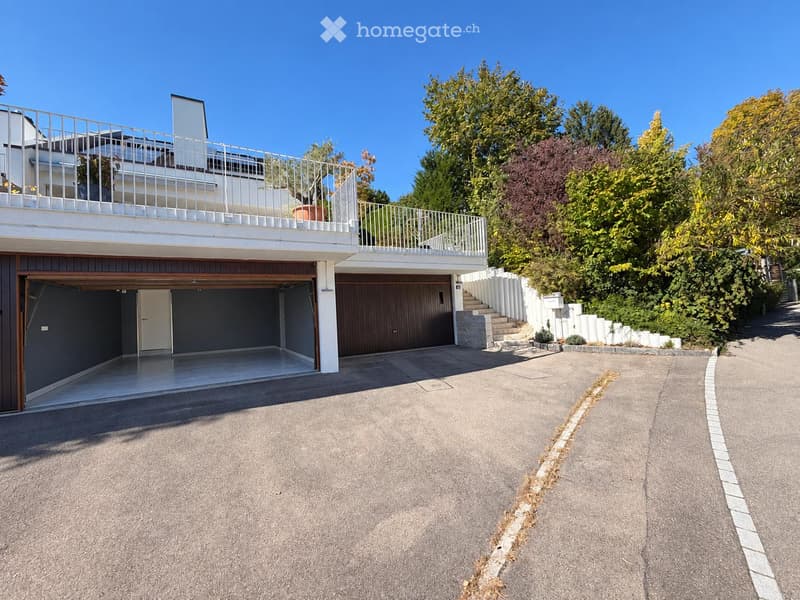 Two-story house with a garage, balcony, and steps leading up to the entrance, surrounded by greenery.