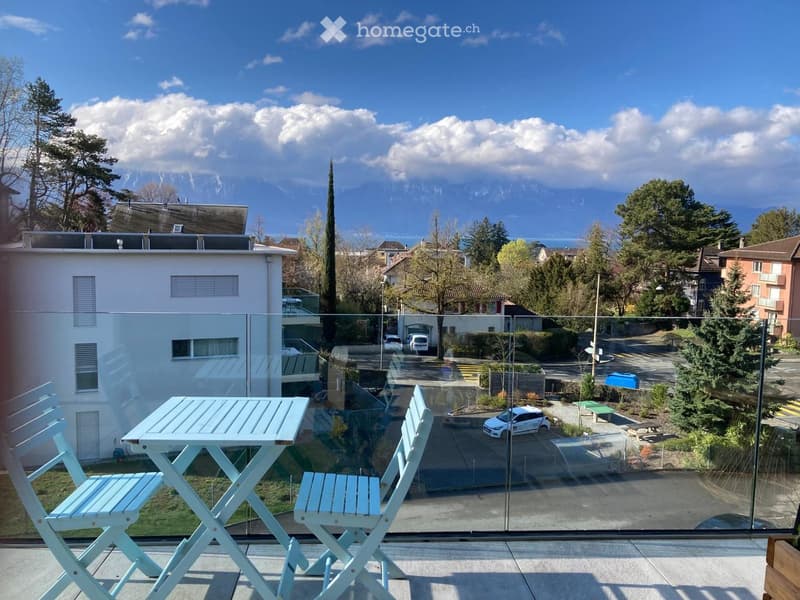 Balcony with a glass railing, a small table, and two chairs, overlooking a scenic view of mountains and a town.