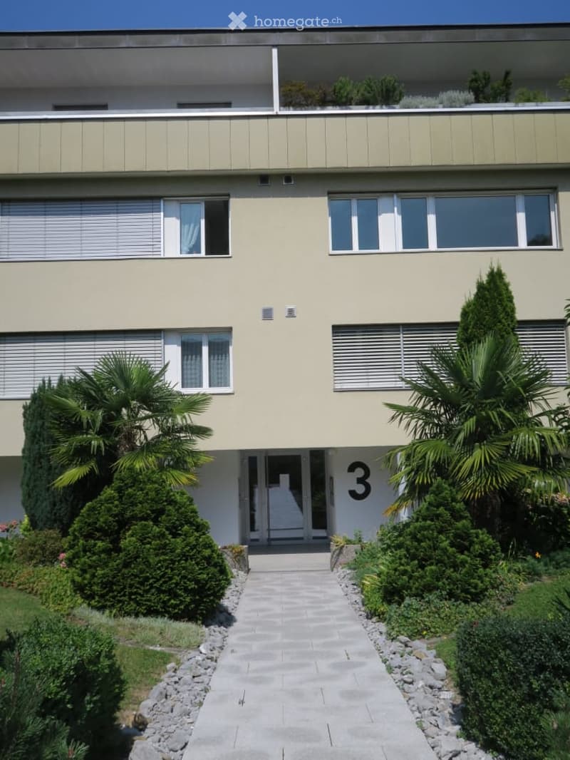 Two-story building with light yellow facade, multiple windows with blinds, and a small garden area with palm trees and shrubs.
