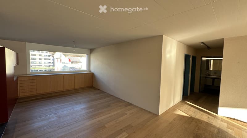 Empty room with wooden flooring, light-colored walls, and built-in cabinets along one wall. A large window provides natural light, and a small adjacent bathroom is visible.