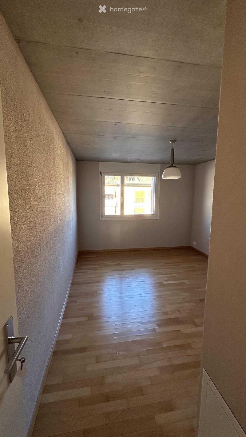 Empty hallway with wooden flooring, white walls, and a single window at the end. A pendant light hangs from the ceiling, and a door is visible on the left side.