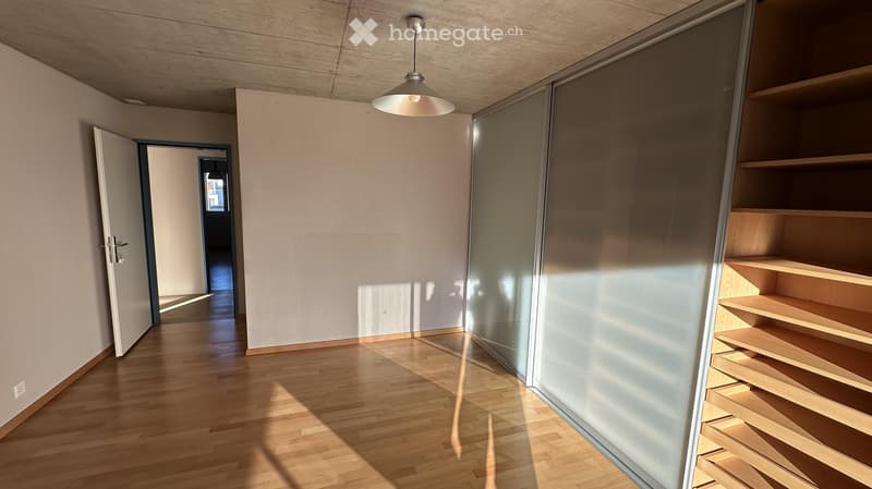 Empty room with wooden flooring, white walls, and built-in wooden shelving unit. A pendant light hangs from the ceiling, and an open doorway leads to another room.