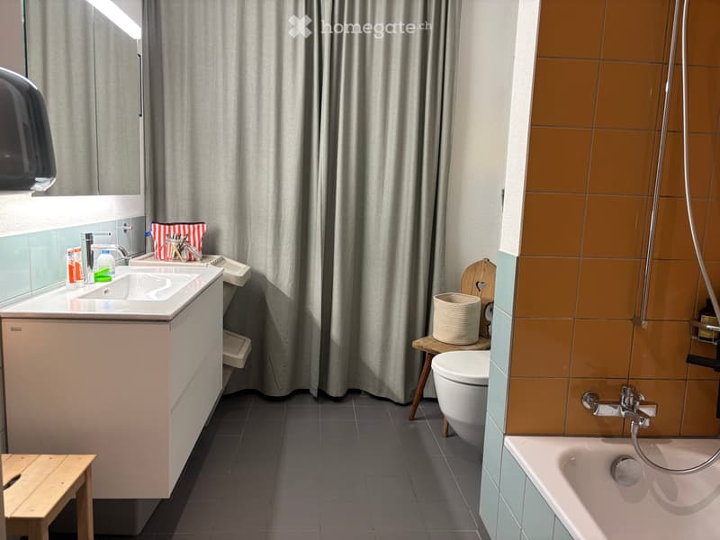 Modern bathroom with a white sink, toilet, bathtub, and wooden stool holding a woven basket. Grey tiles on the floor and walls, with a large window and grey curtain.