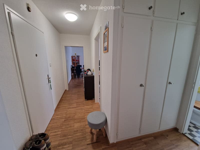A narrow hallway with wooden flooring, white walls, and several doors. A small grey stool is placed near the center, and a pair of shoes is on the floor to the left. A framed picture hangs on the wall to the right, and a room with a desk and chairs is vis