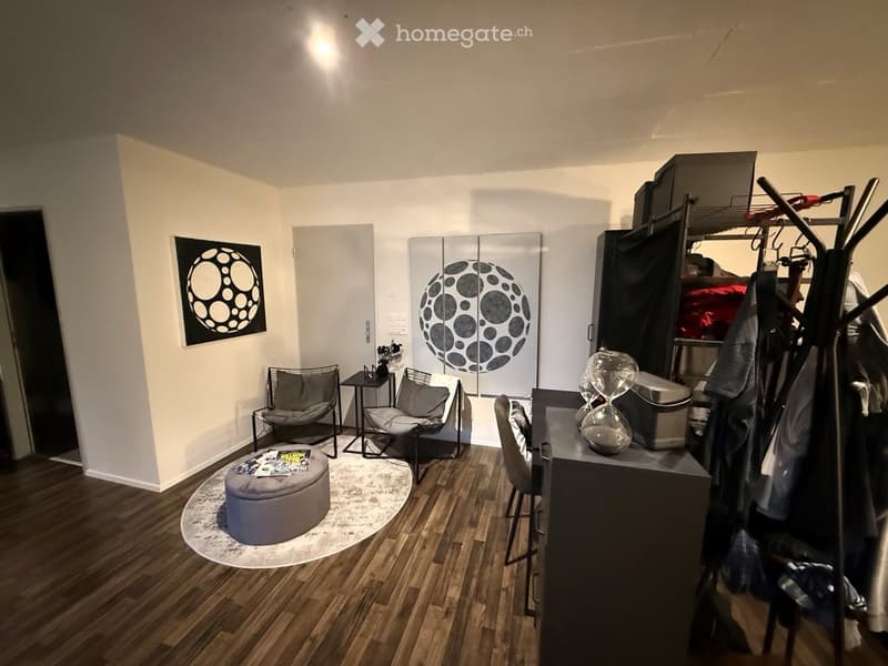 A modern, well-lit room with wooden flooring, a round grey stool, two chairs, a small table, a mirror, and artwork on the wall.