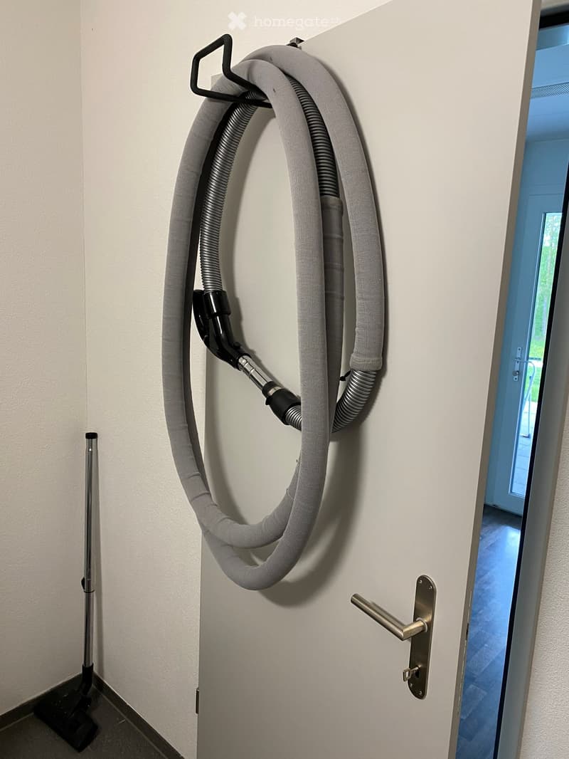 A hallway with a vacuum cleaner hose mounted on the wall and a door leading to another room.