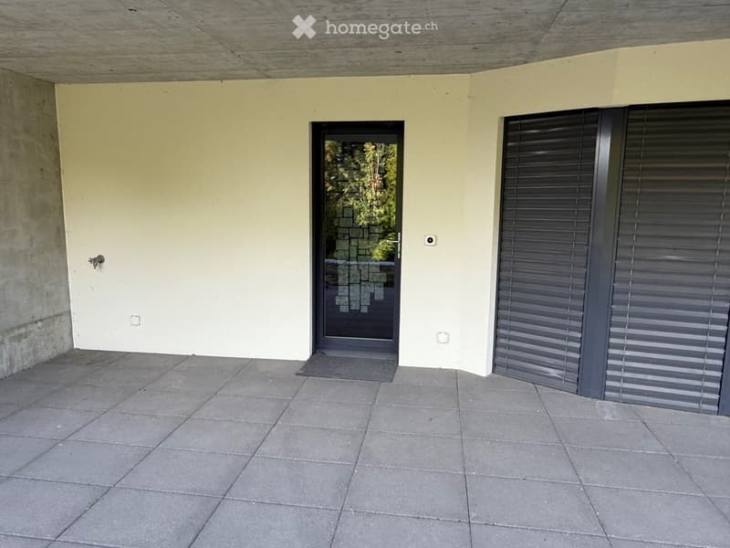 A modern outdoor patio with a tiled floor, a glass door leading outside, and a closed metal shutter on the right side.