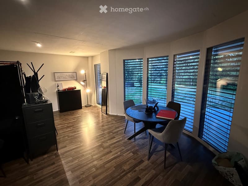 Modern dining area with wooden flooring, large windows with blinds, round table with chairs, cabinet, mirror, lamp, and decorative items.