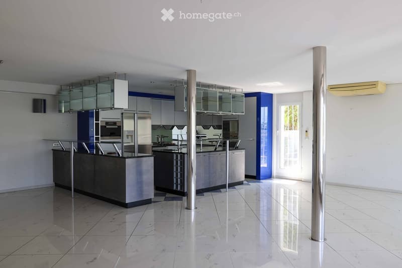 Modern kitchen with stainless steel appliances, glass-fronted cabinets, and a central island. The space features a polished tile floor and large windows allowing natural light.