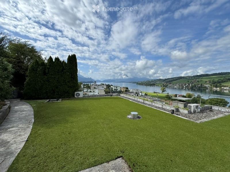 Lush green garden with a panoramic view of a lake and distant mountains under a partly cloudy sky.