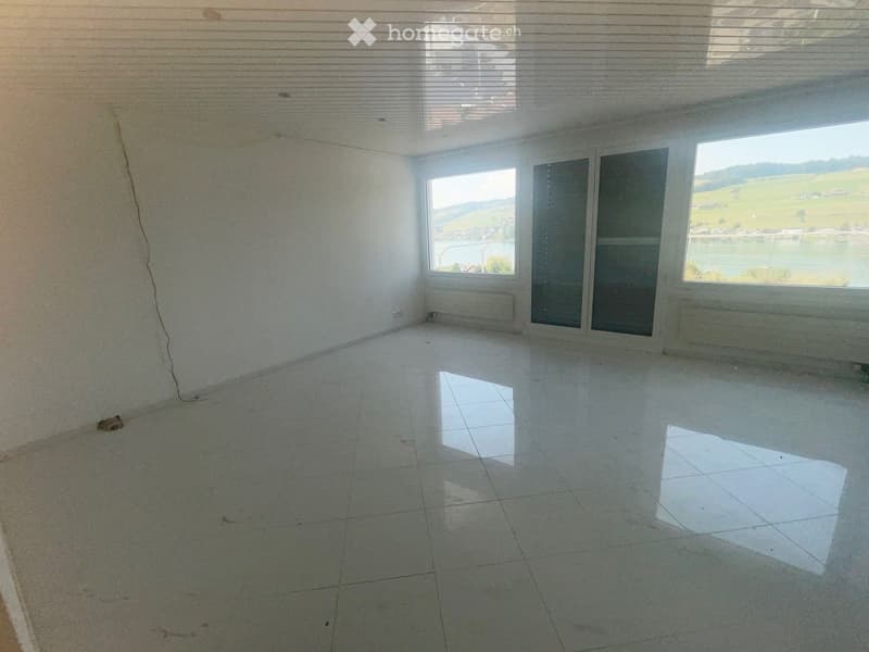Empty room with white tiled floor, large windows, and a scenic view of a lake and green landscape.