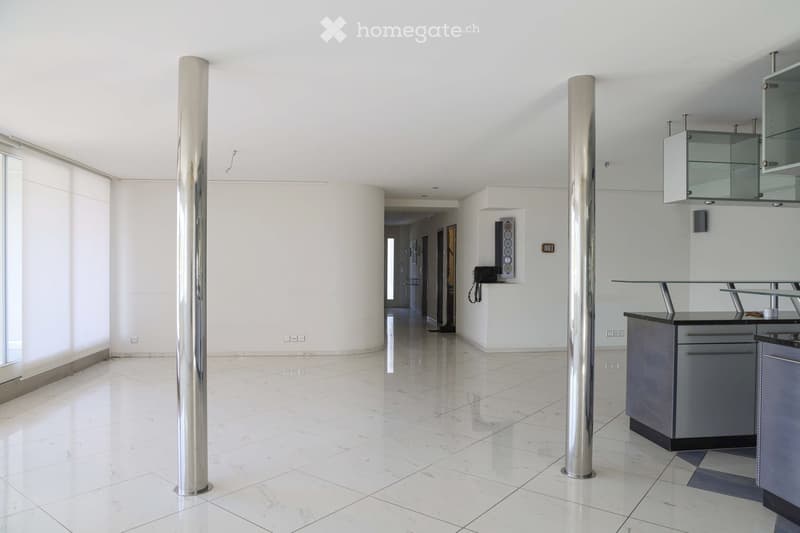 Open, modern space with white walls, polished marble floors, and large glass windows. Minimalist design with stainless steel columns and a small kitchen area featuring grey cabinets and a glass countertop.