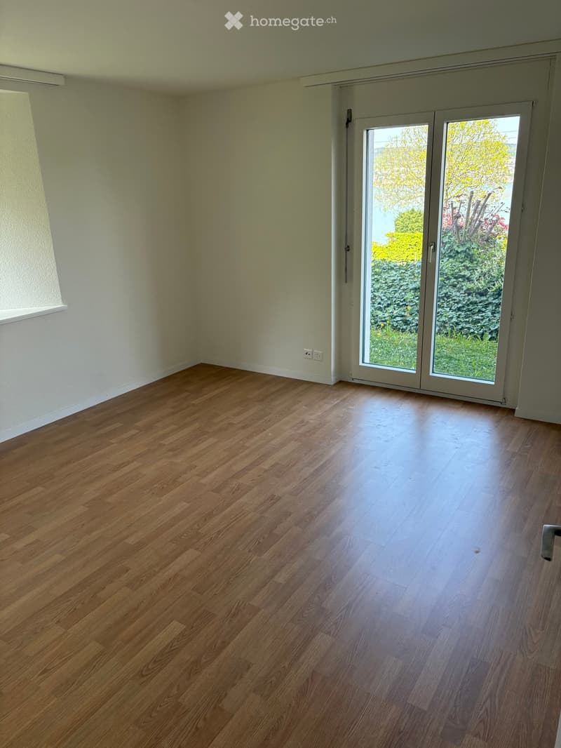 Empty room with wooden flooring, white walls, and large sliding glass doors leading to a garden area.