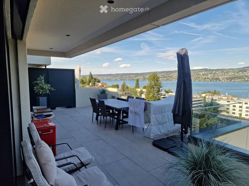 Modern balcony with tiled floor, outdoor dining set, lounge chairs, umbrella, and scenic lake view.
