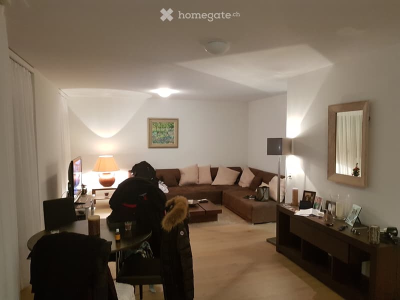 Living room with a brown sectional sofa, coffee table, side table, lamp, mirror, and television. Wooden flooring and white walls.