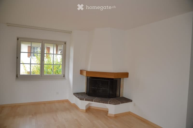 Empty room with light wooden flooring, white walls, a fireplace with a wooden mantel, and a window with a view of greenery outside.