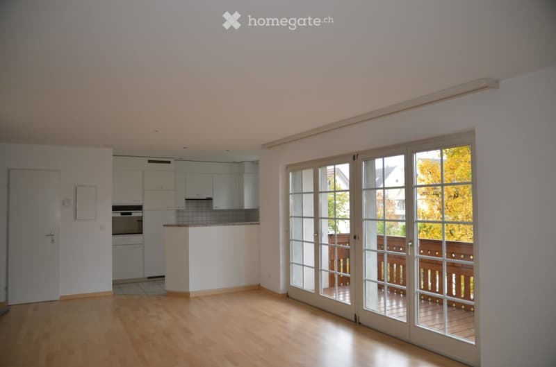 Bright, unfurnished room with wooden flooring, large glass sliding doors leading to a balcony, and a small kitchen area with white cabinets and appliances.