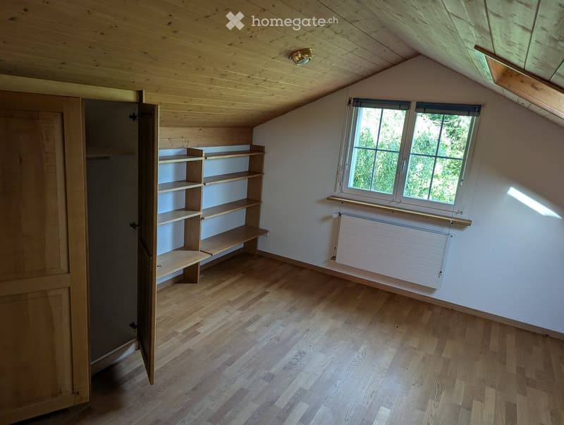 Empty attic room with wooden floors, slanted wooden ceiling, built-in wooden shelves, and a window with a radiator below.