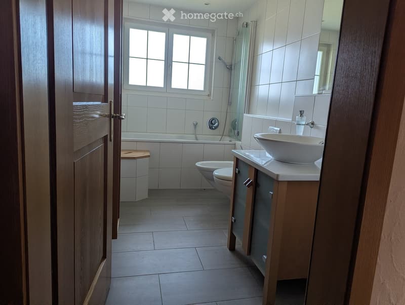 Modern bathroom with white tiles, a bathtub, toilet, sink, and a glass-enclosed shower area. Features a wooden door and a window for natural light.