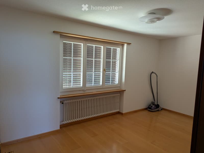 Empty room with wooden flooring, white walls, large window with blinds, radiator, and a vacuum cleaner in the corner.