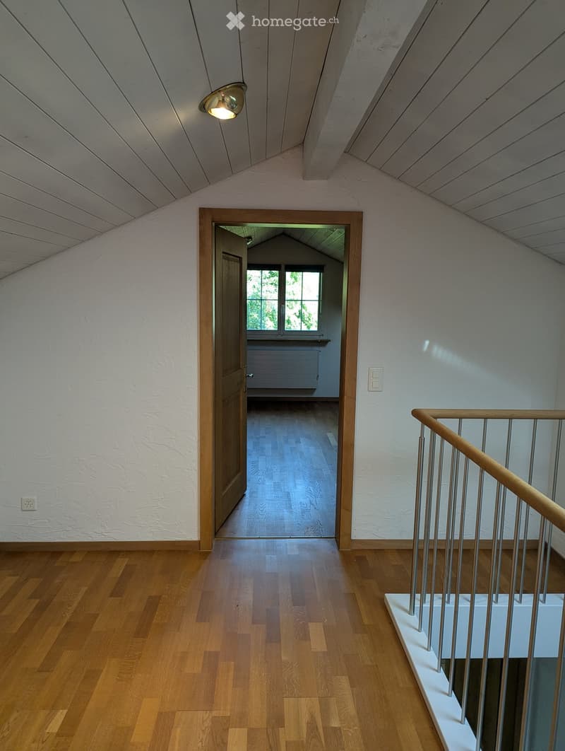Bright hallway with wooden flooring, white walls, and a staircase leading upwards. A door opens to another room with large windows and wooden flooring.