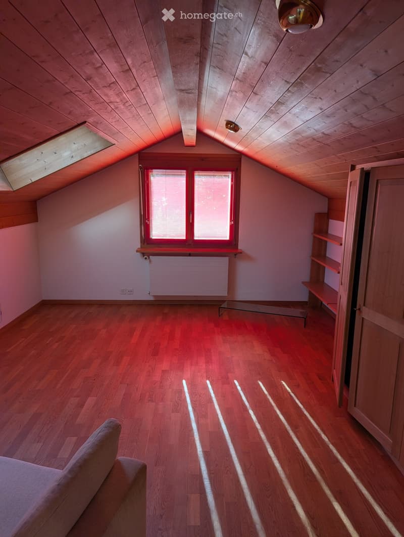 Spacious attic room with wooden floors, slanted wooden ceiling, large windows, built-in shelves, and a sofa.